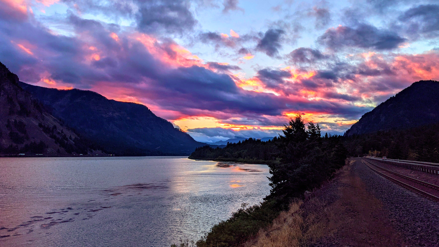 Pictured: A photograph of the Columbia River Gorge at sunset. The river runs through the center of the photo and into the horizon where the sky is painted with blue, pink, orange and purple clouds.