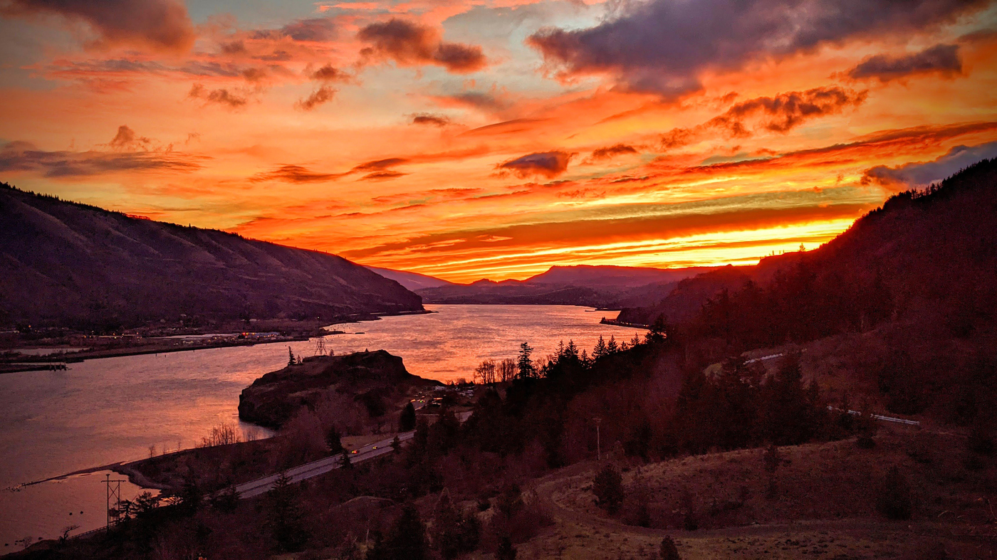 Pictured: A photo of the Columbia River Gorge at sunset. A silver river cuts deep into the landscape and reflects a orange, blue and yellow sky.