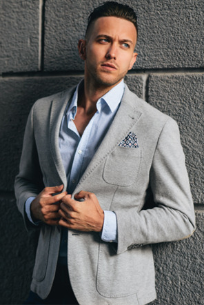 Professional man wearing JC Broadway Collection light grey cotton blazer with patterned pocket square and blue dress shirt, posed against industrial concrete wall