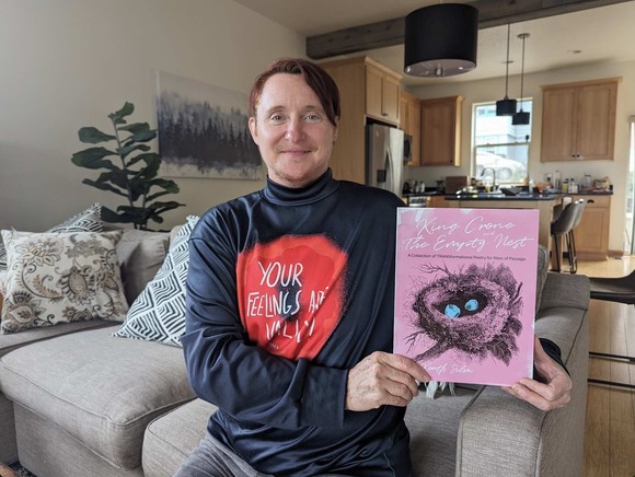 Keath Silva poses with his second book of poetry, King Crone and the Empty Nest. He wears a shirt designed by Mars Wright, who makes clothing for nonbinary and trans people. The shirt reads "Your feelings are valid."